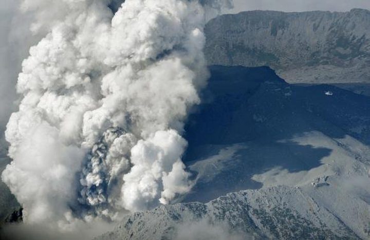 A helicopter (R top) flies near rising smoke and mountain lodges (R bottom) covered with volcanic...